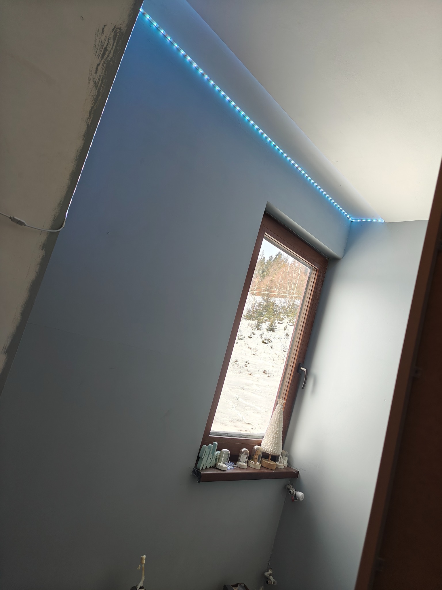 Poddasze z oknem w drewnianej ramie, oświetlone listwą LED. Ściany pomalowane na szaro, na parapecie dekoracje. Widok na zaśnieżone drzewa.