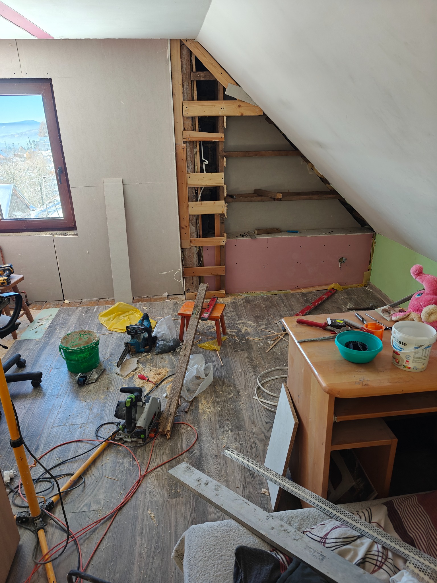 Poddasze w trakcie adaptacji: widoczne płyty karton-gips, drewniana konstrukcja, narzędzia i bałagan. Okno z widokiem na zimowy krajobraz.