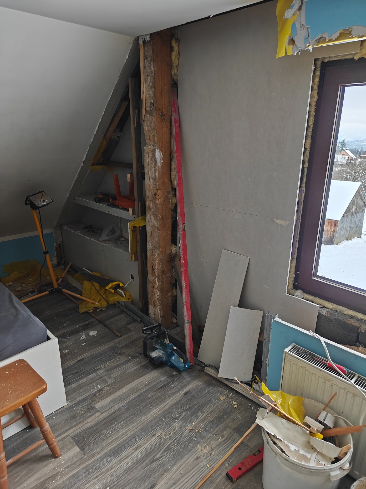 Poddasze w trakcie remontu: odsłonięta konstrukcja, płyty karton-gips, narzędzia i lampa budowlana. Widok na zaśnieżony krajobraz przez okno.