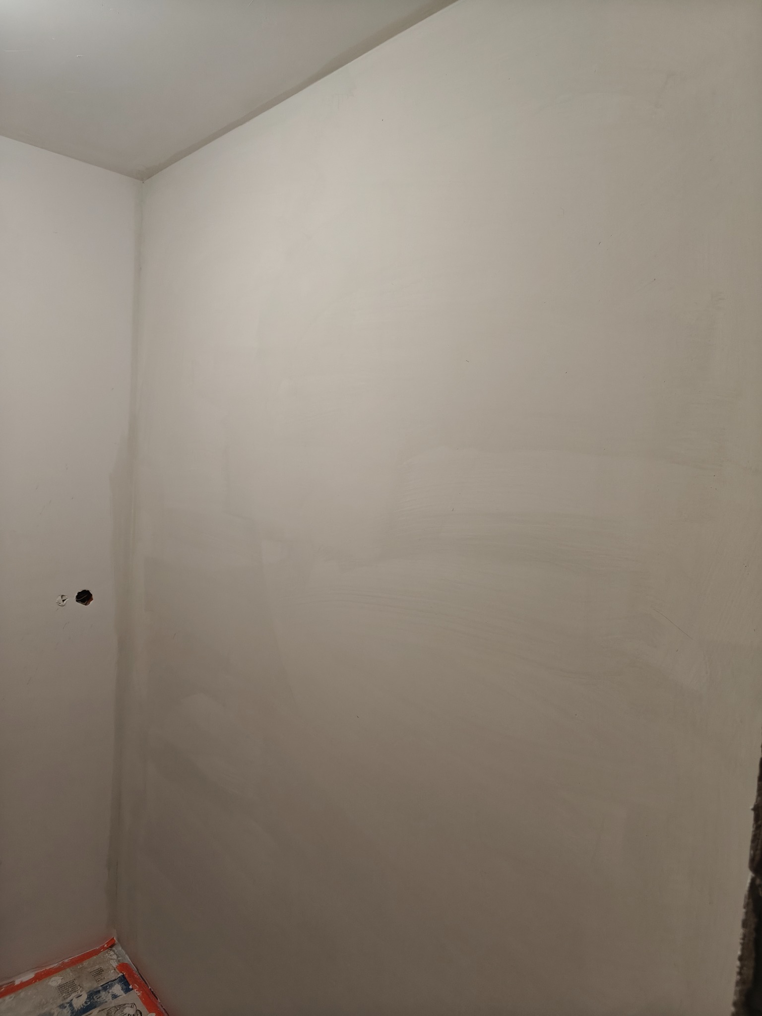 Świeżo wyszpachlowana ściana w rogu pomieszczenia, widoczne ślady po szpachlowaniu, przygotowana do dalszych prac wykończeniowych. Otwór na gniazdko elektryczne.