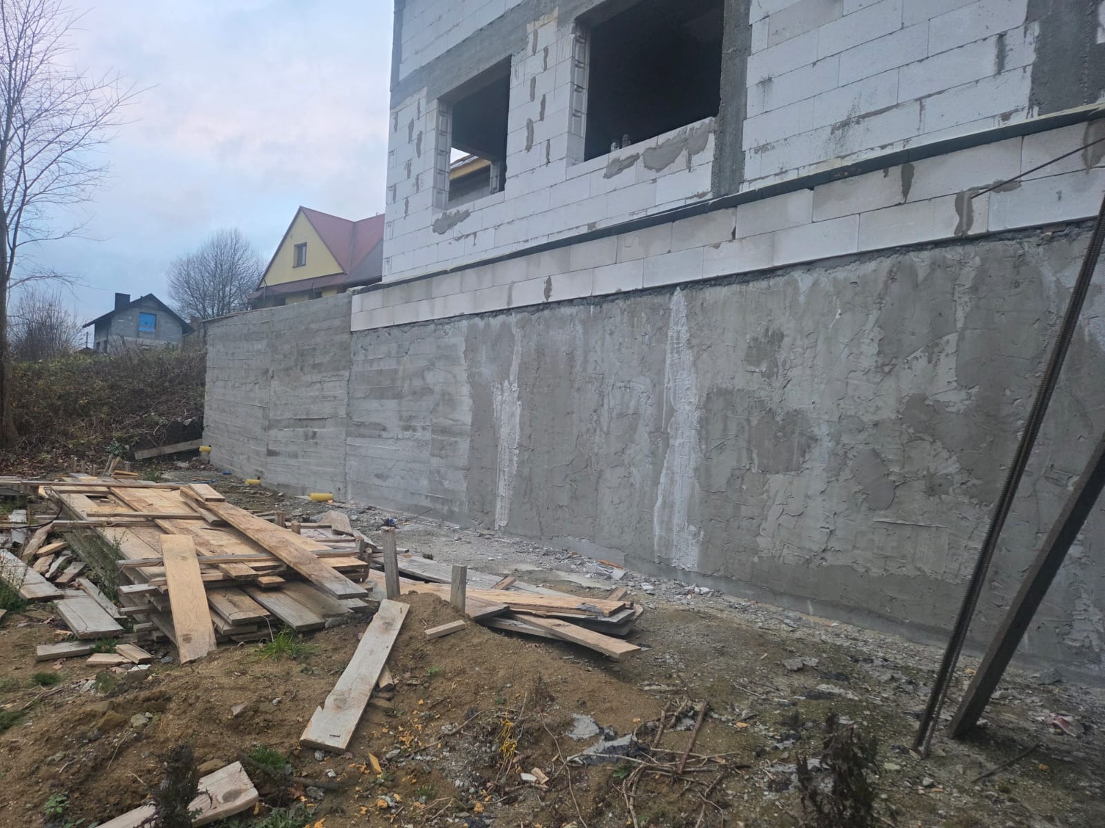 Surowy stan domu z betonu komórkowego, widoczny fundament z szorstką fakturą i stertą desek w pobliżu. Budowa w toku, zarys domów w tle.