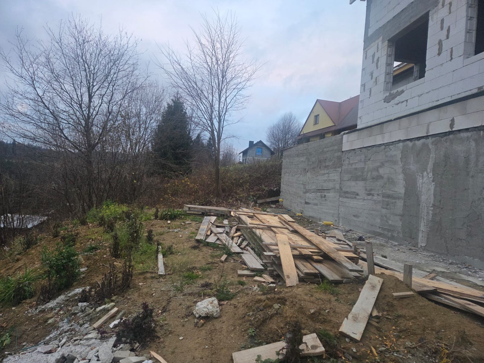 Surowy stan domu murowanego w trakcie budowy, widoczne fundamenty i ściany z bloczków, drewniane deski na ziemi, drzewa bez liści w tle, pochmurne niebo.