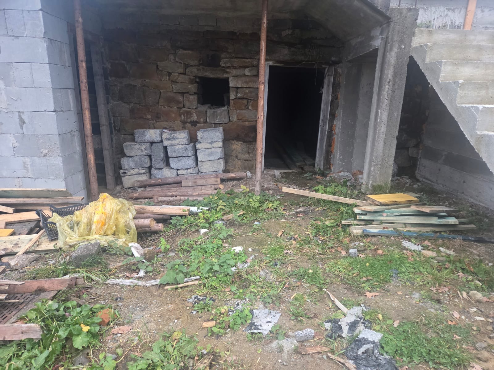 Surowy stan budynku w trakcie budowy: kamienne ściany, betonowe elementy, materiały budowlane na ziemi. Widoczne niedokończone schody i otwór wejściowy.