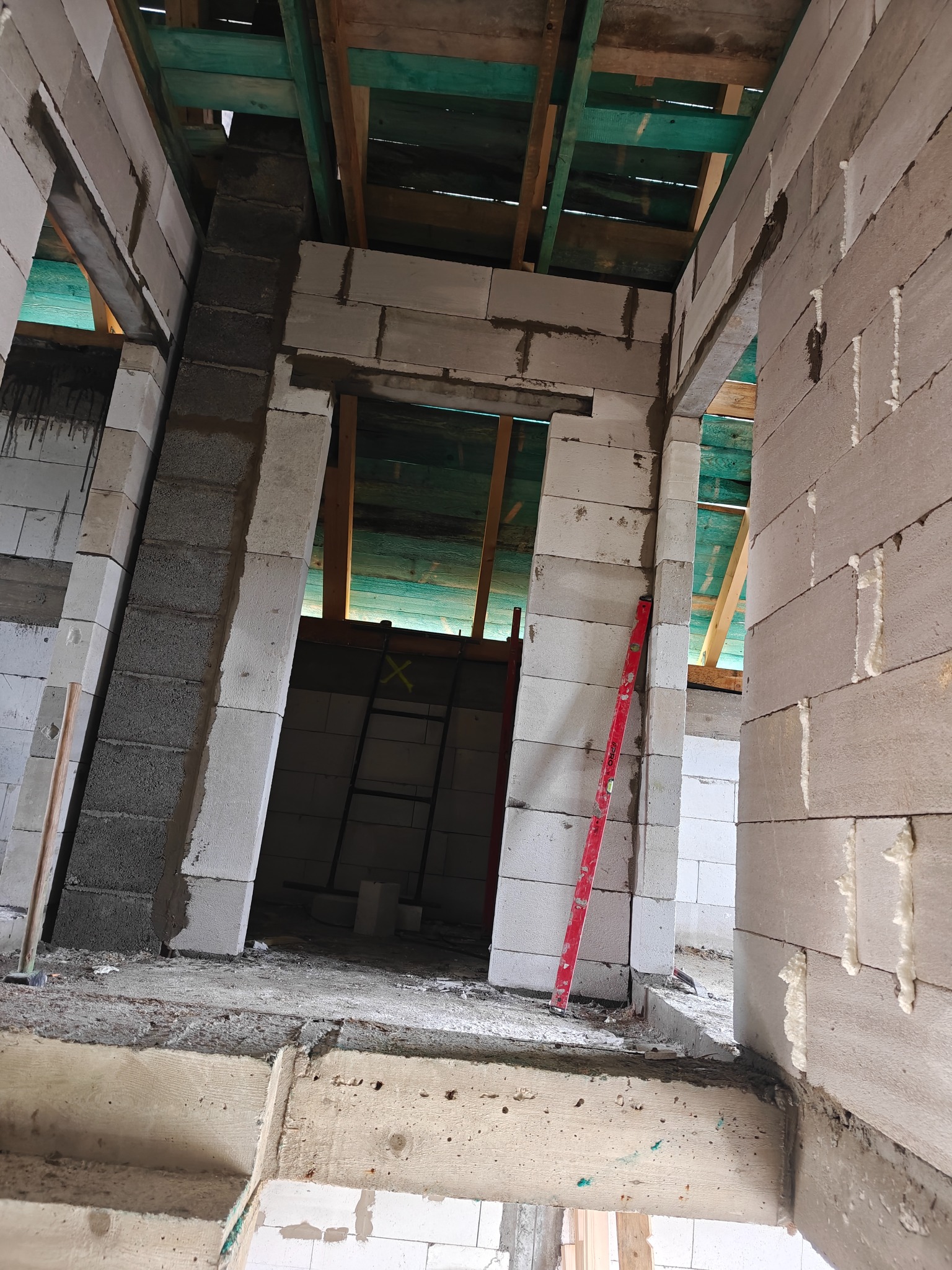 Wnętrze w trakcie budowy z białych bloczków, widoczny poziomica oparta o ścianę, surowe drewniane elementy konstrukcyjne stropu i ślady zaprawy murarskiej.