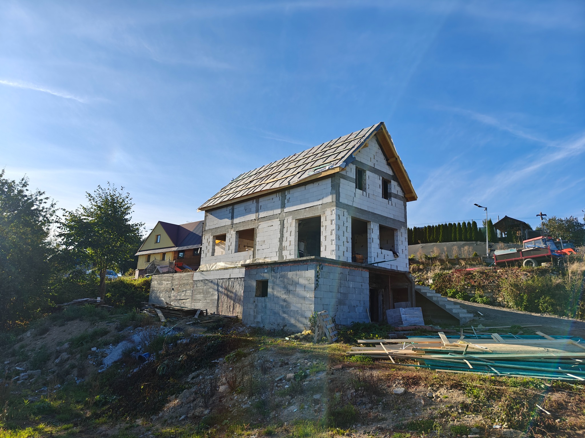 Surowy stan domu z białego bloczka, częściowo zadaszony, na wzgórzu w słoneczny dzień. Widoczne materiały budowlane i otoczenie w trakcie budowy.