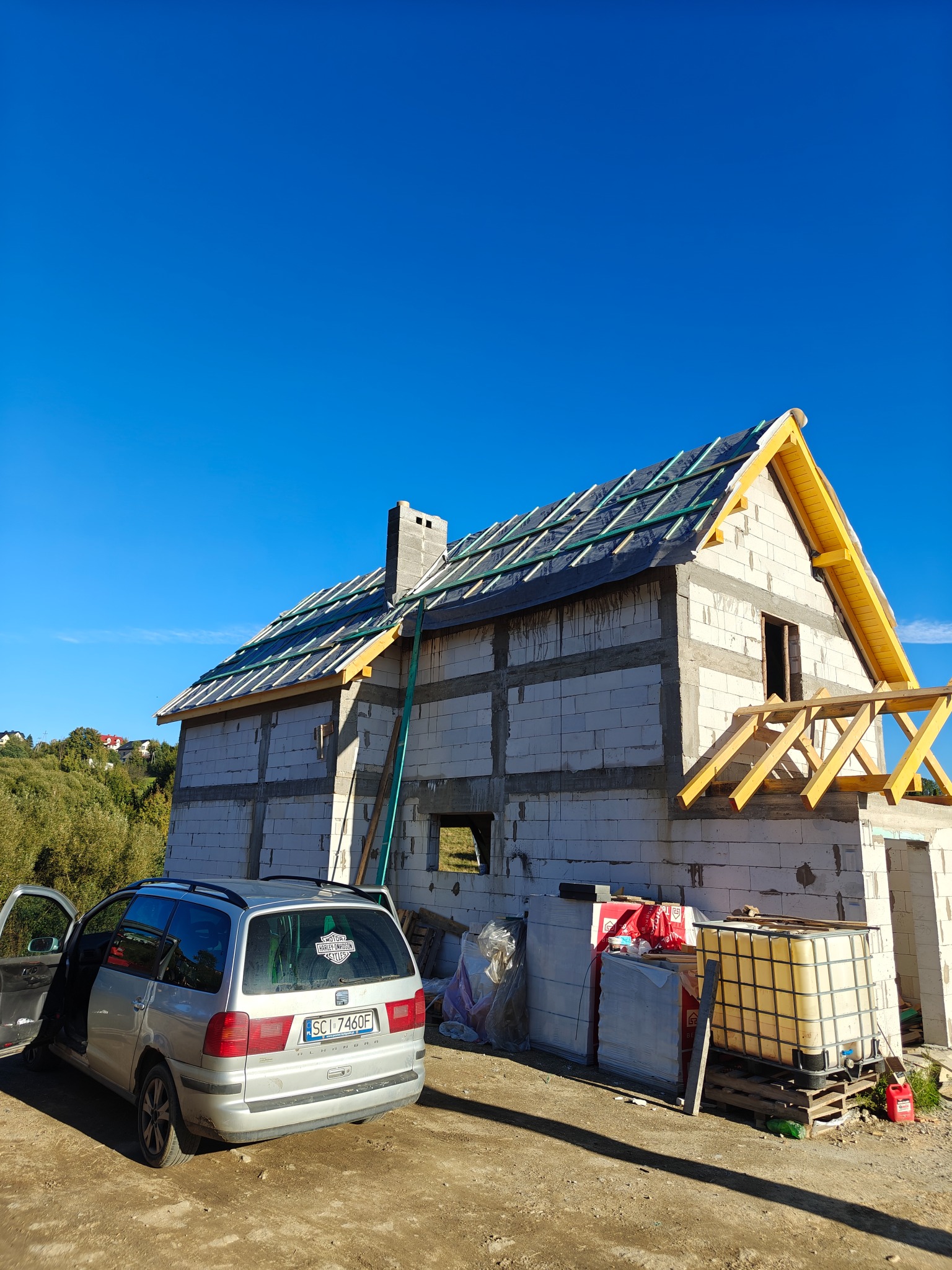 Budowa domu z białego bloczka: surowy stan, widoczne elementy konstrukcji dachu, rusztowania. Na pierwszym planie srebrny samochód osobowy.