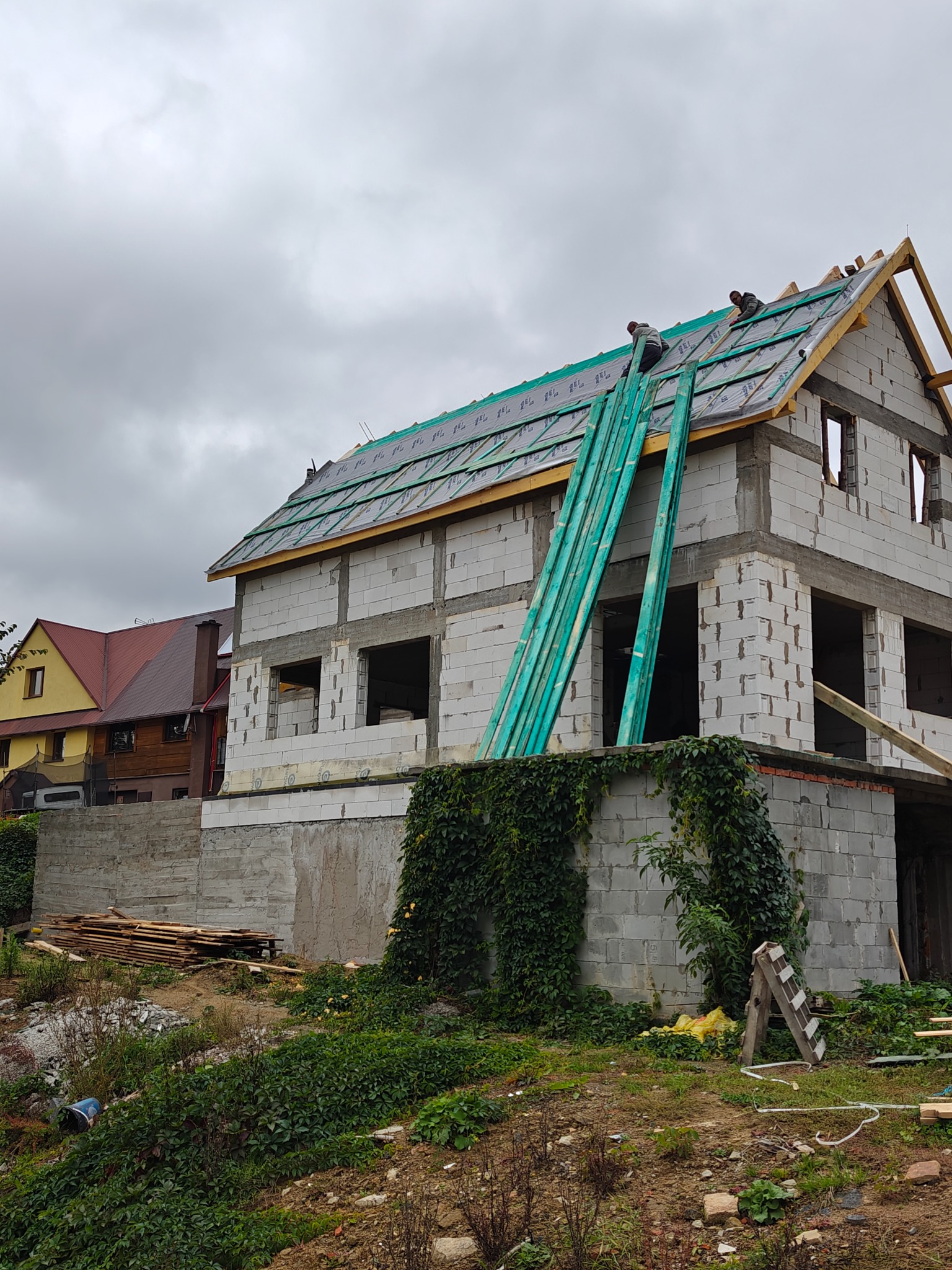 Budowa domu z białych bloczków: ekipa montuje izolację dachu, zielone deski opierają się o ścianę, w tle sąsiedni dom i pochmurne niebo.