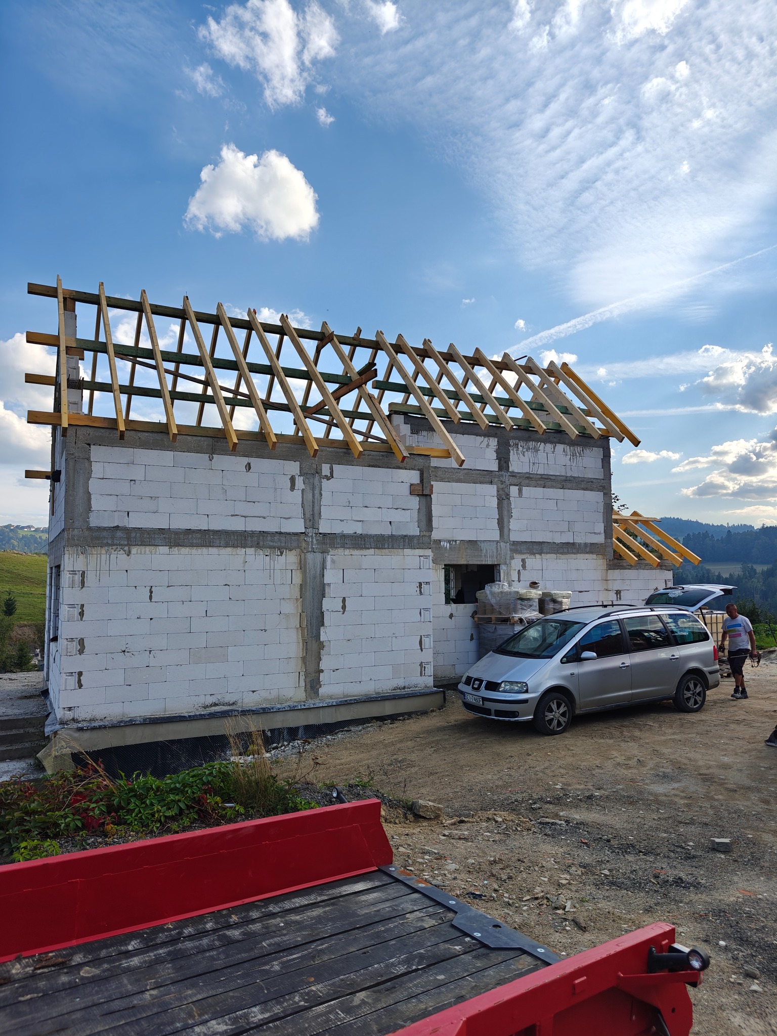 Budowa domu z białych bloczków, widoczne drewniane elementy konstrukcji dachu, auto dostawcze z materiałami budowlanymi w tle, krajobraz górski.