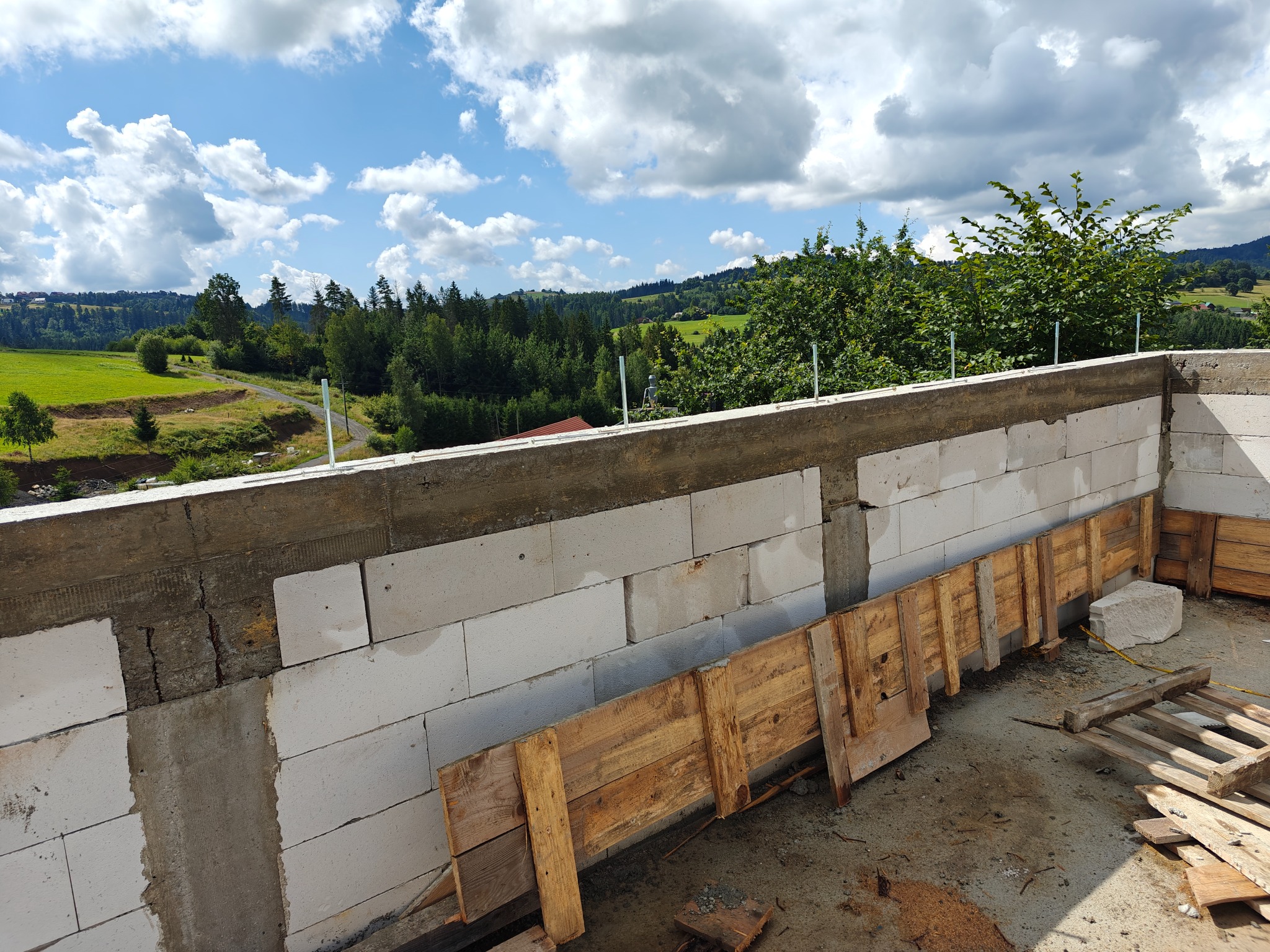 Wznoszenie ściany z betonu komórkowego Ytong z drewnianym szalunkiem na tle malowniczego krajobrazu z zielonymi wzgórzami i błękitnym niebem z chmurami.