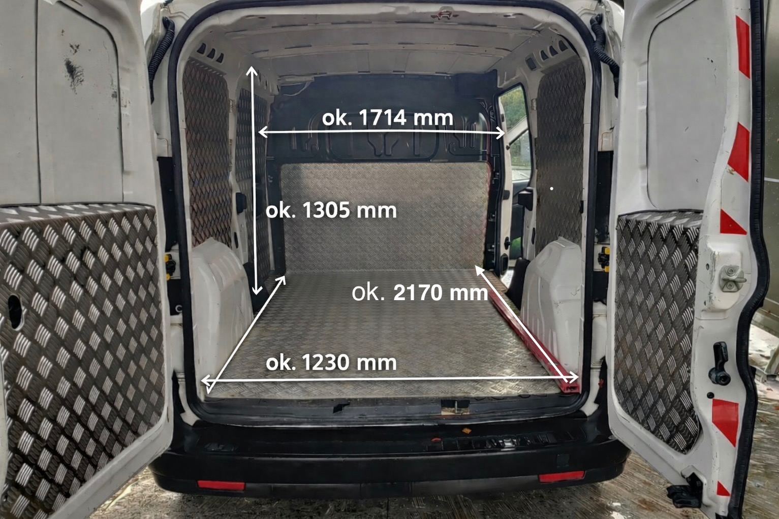 Wnętrze białego busa z otwartymi drzwiami, widok z tyłu. Podłoga i ściany zabezpieczone blachą ryflowaną. Na zdjęciu wymiary przestrzeni ładunkowej.