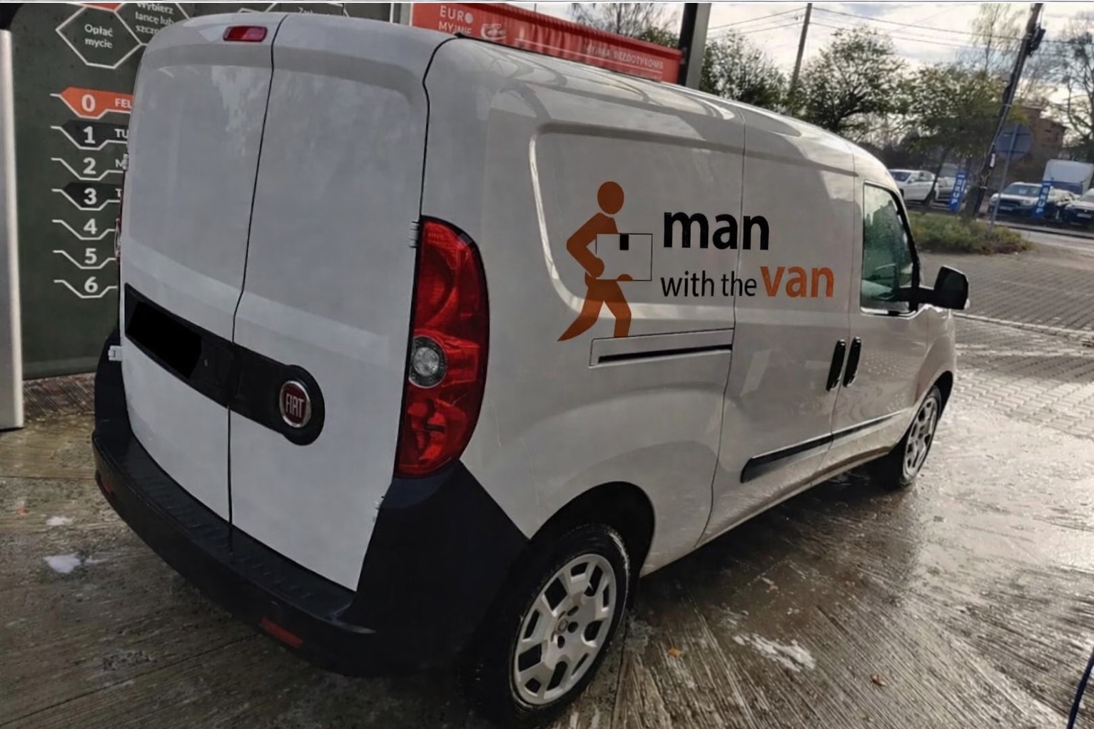 Biały Fiat Doblo z logo 'man with the van' po myciu, widok z tyłu pod kątem. Ślady wody na ziemi, w tle myjnia samochodowa i inne pojazdy.