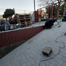 delta remont - Układanie cegiełek na murku przy posesji. Widoczne palety z workami, narzędzia i osoba w trakcie pracy. Cegiełki w kolorze czerwonym, ułożone w regularny wzór.