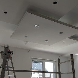 delta remont - Biały sufit podwieszany z zamontowanymi punktowymi lampami LED, widok z dołu, drabiny malarskie w pomieszczeniu, okno w tle.