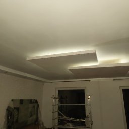 delta remont - Nowoczesny sufit podwieszany z oświetleniem LED w trakcie montażu, widoczna drabina i zabezpieczone meble folią malarską. Neutralna kolorystyka wnętrza.