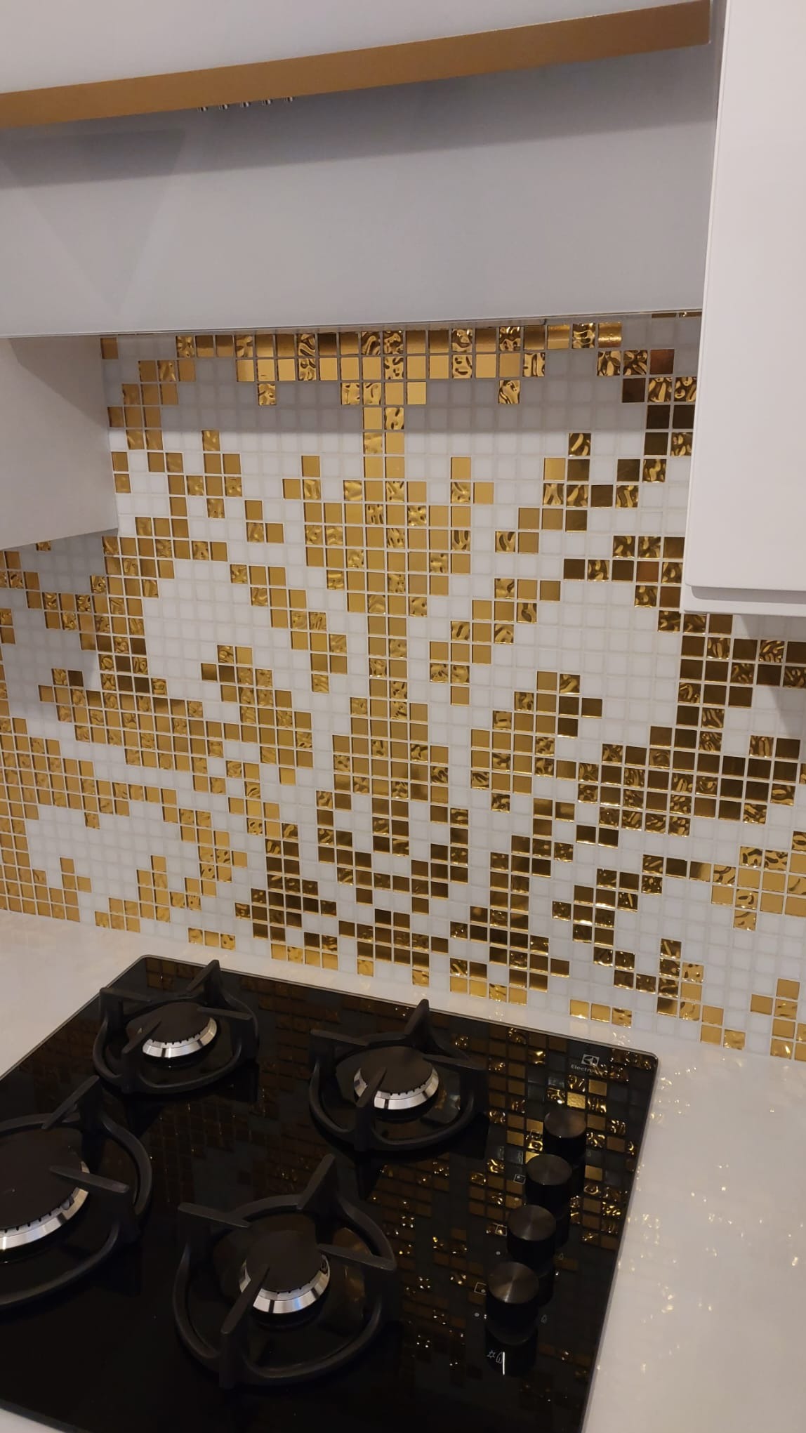 Błyszcząca mozaika w odcieniach złota i bieli zdobi ścianę nad czarną płytą gazową w nowoczesnej kuchni. Uwagę przykuwa wzór mozaiki przypominający rozproszone konfetti.
