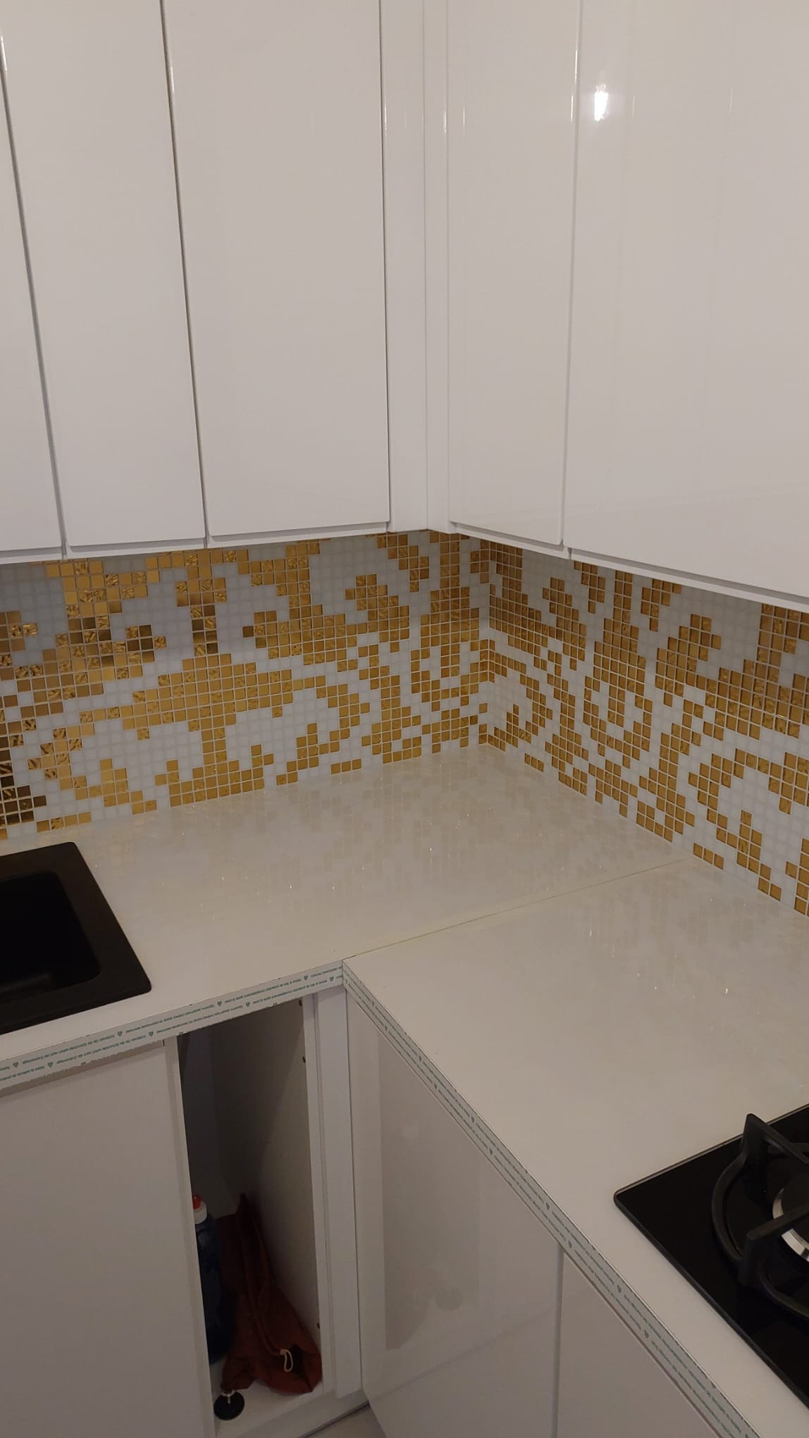 Biała, błyszcząca kuchnia z mozaiką w odcieniach złota i bieli nad blatem. Widoczne szafki górne i fragment płyty grzewczej. Nowoczesny design.