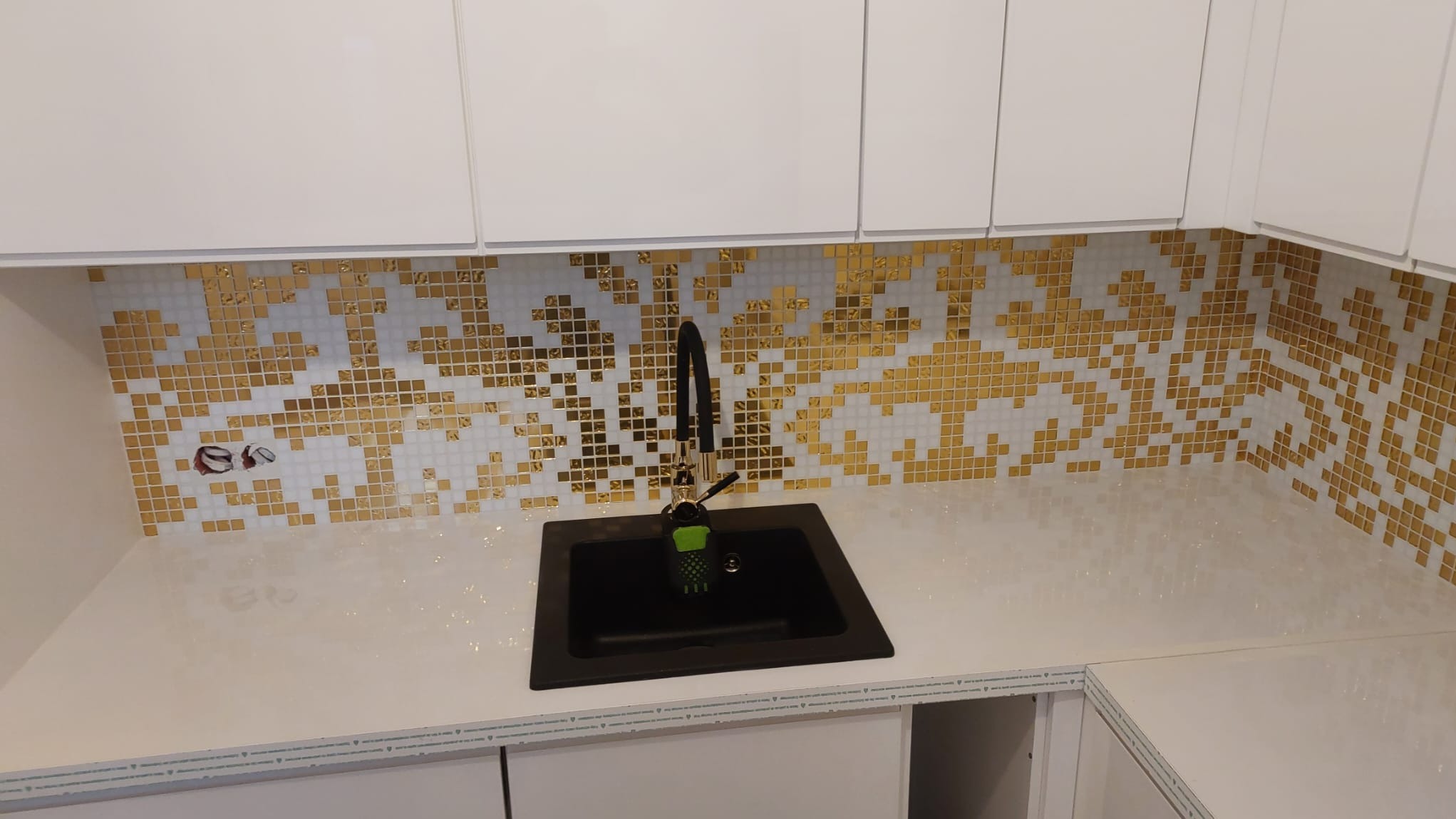 Błyszcząca mozaika w odcieniach złota i bieli zdobi ścianę nad czarnym zlewem w nowoczesnej kuchni z białymi szafkami. Widoczne gniazdko elektryczne.