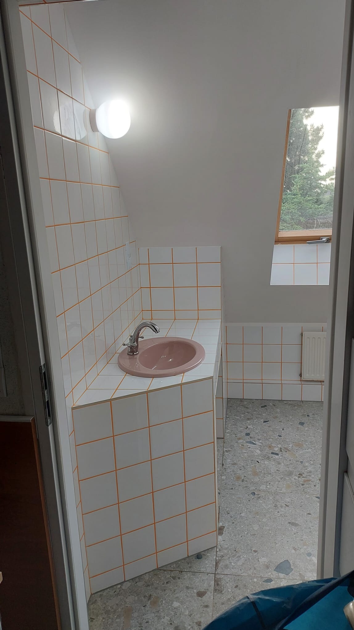 Łazienka na poddaszu z różową umywalką w zabudowie z białych płytek z pomarańczową fugą. Widoczne okno dachowe i grzejnik. Podłoga lastryko.