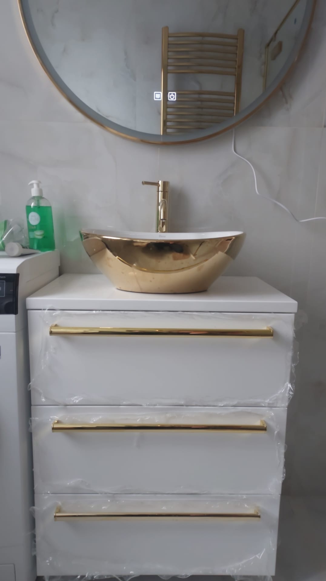 Nowoczesna łazienka: złota umywalka na białej szafce z trzema szufladami, okrągłe lustro z podświetleniem LED i grzejnik drabinkowy w tle.
