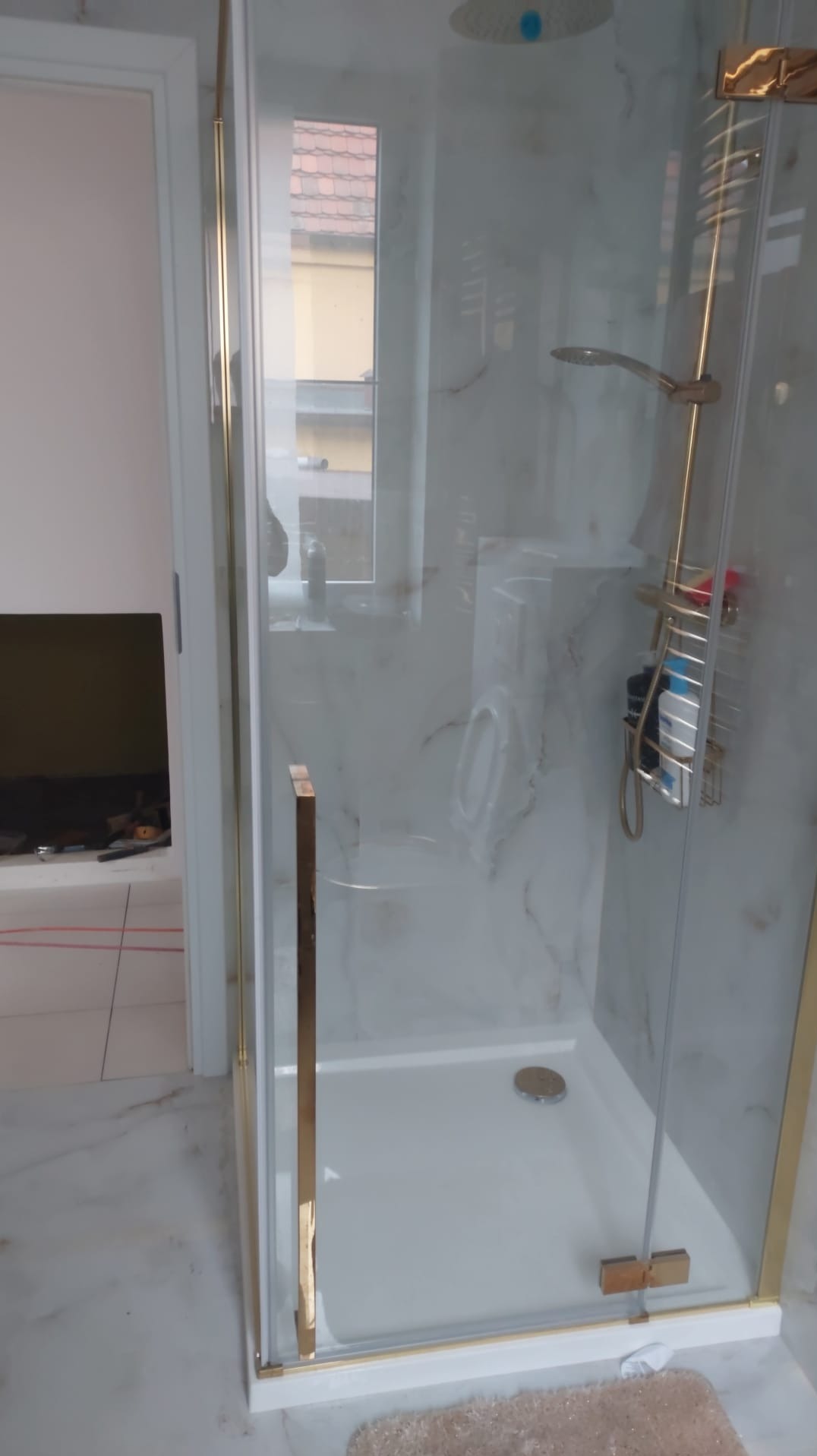 Nowoczesna kabina prysznicowa z złotymi elementami wykończeniowymi i marmurowym wzorem na ścianach, widoczna z boku, z fragmentem łazienki w tle.