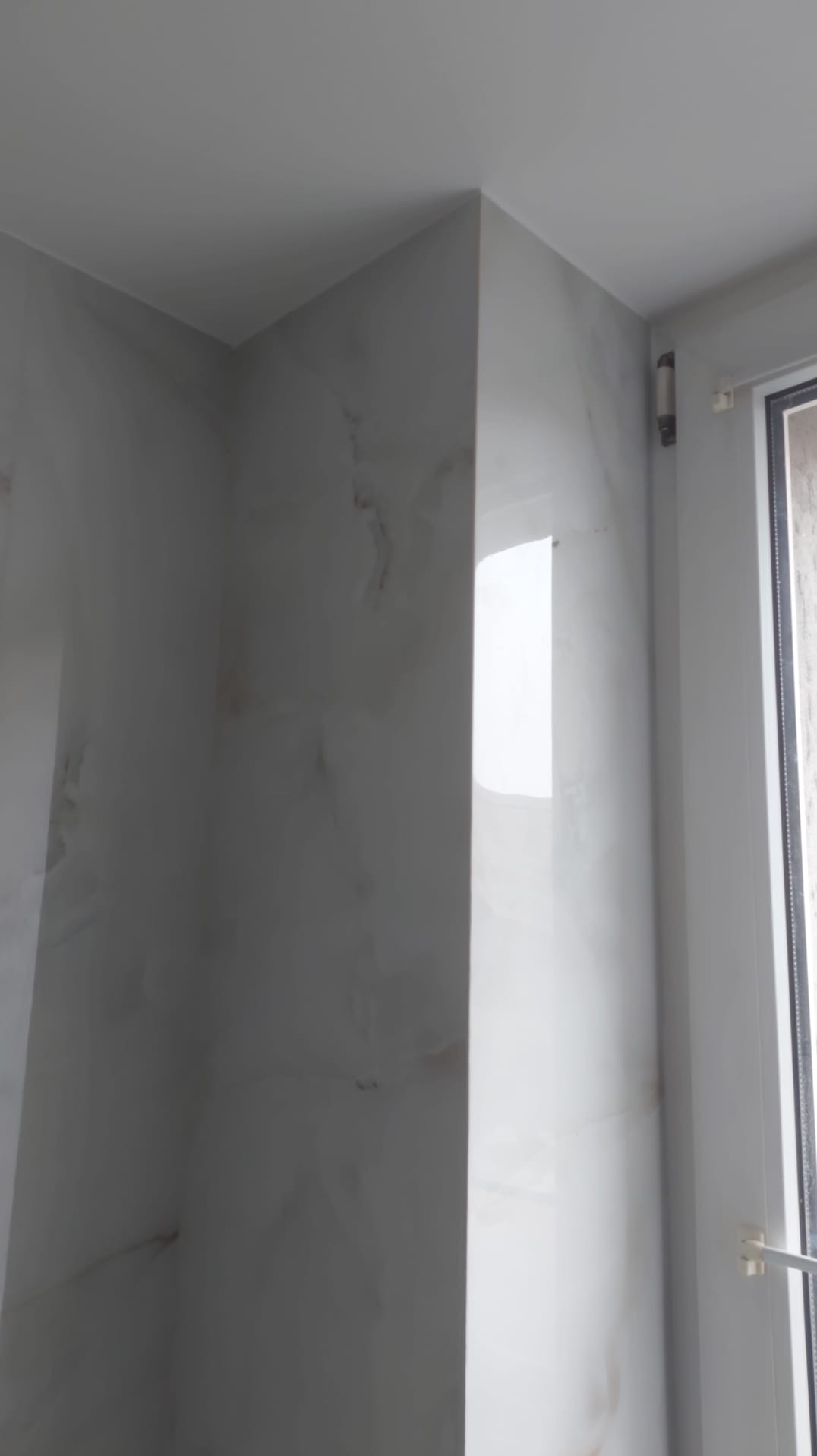 Wykończony narożnik łazienki z białymi, marmurkowymi płytkami, widoczny fragment okna. Minimalistyczny design, jasne wnętrze.