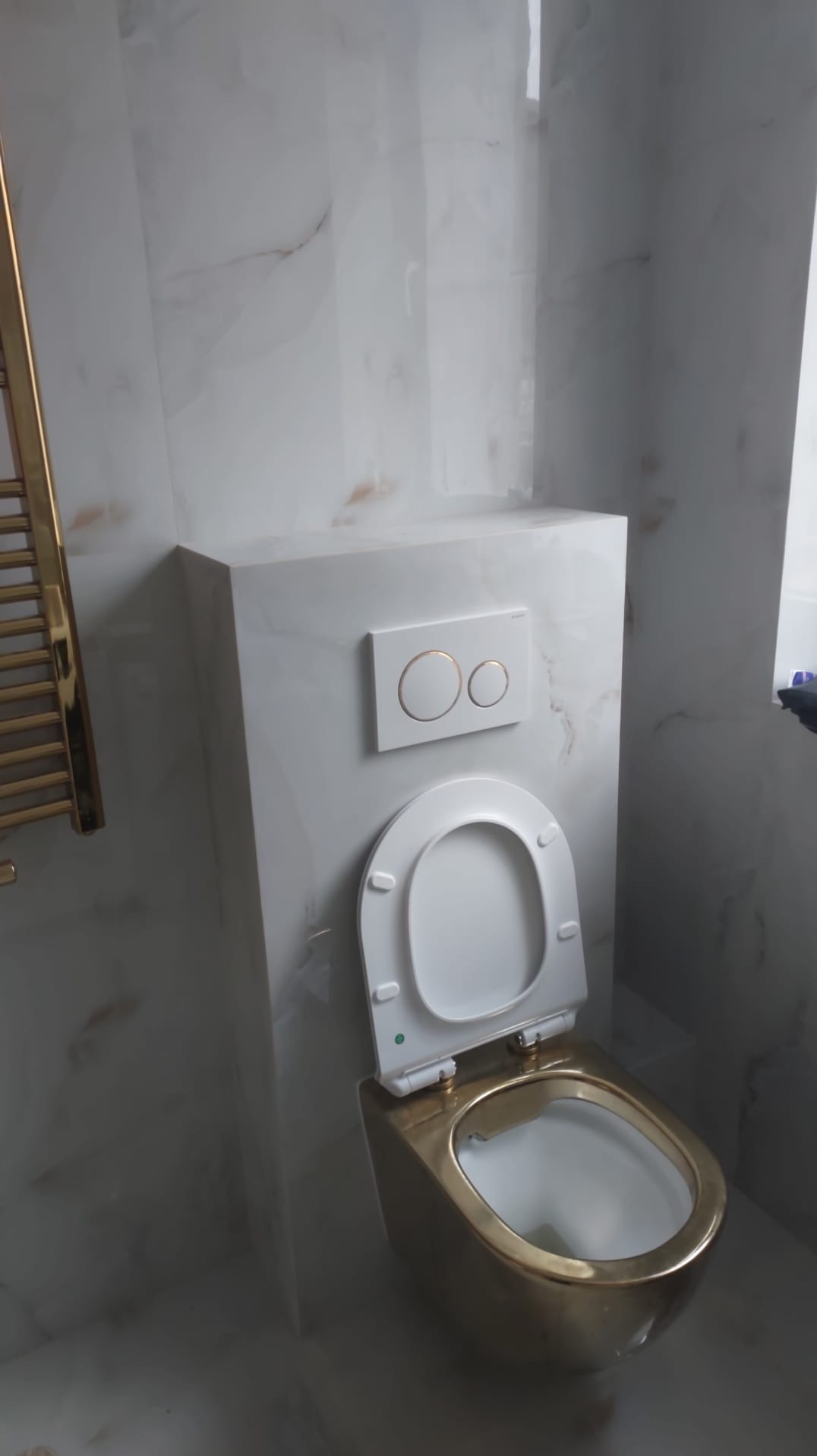 Podwieszana, złota toaleta z białą deską i przyciskiem spłukującym na tle marmurowej ściany w łazience. Widoczny fragment grzejnika.