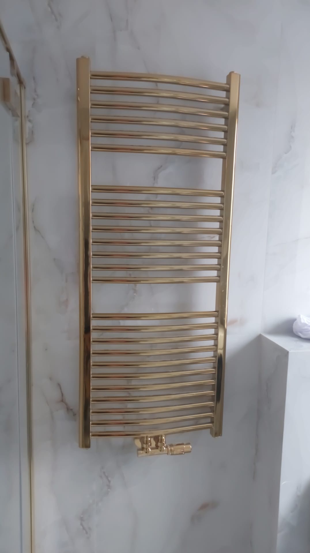 Złoty, dekoracyjny grzejnik łazienkowy na ścianie z marmurowym wzorem. Widoczne zawory i fragment kabiny prysznicowej. Eleganckie wykończenie łazienki.