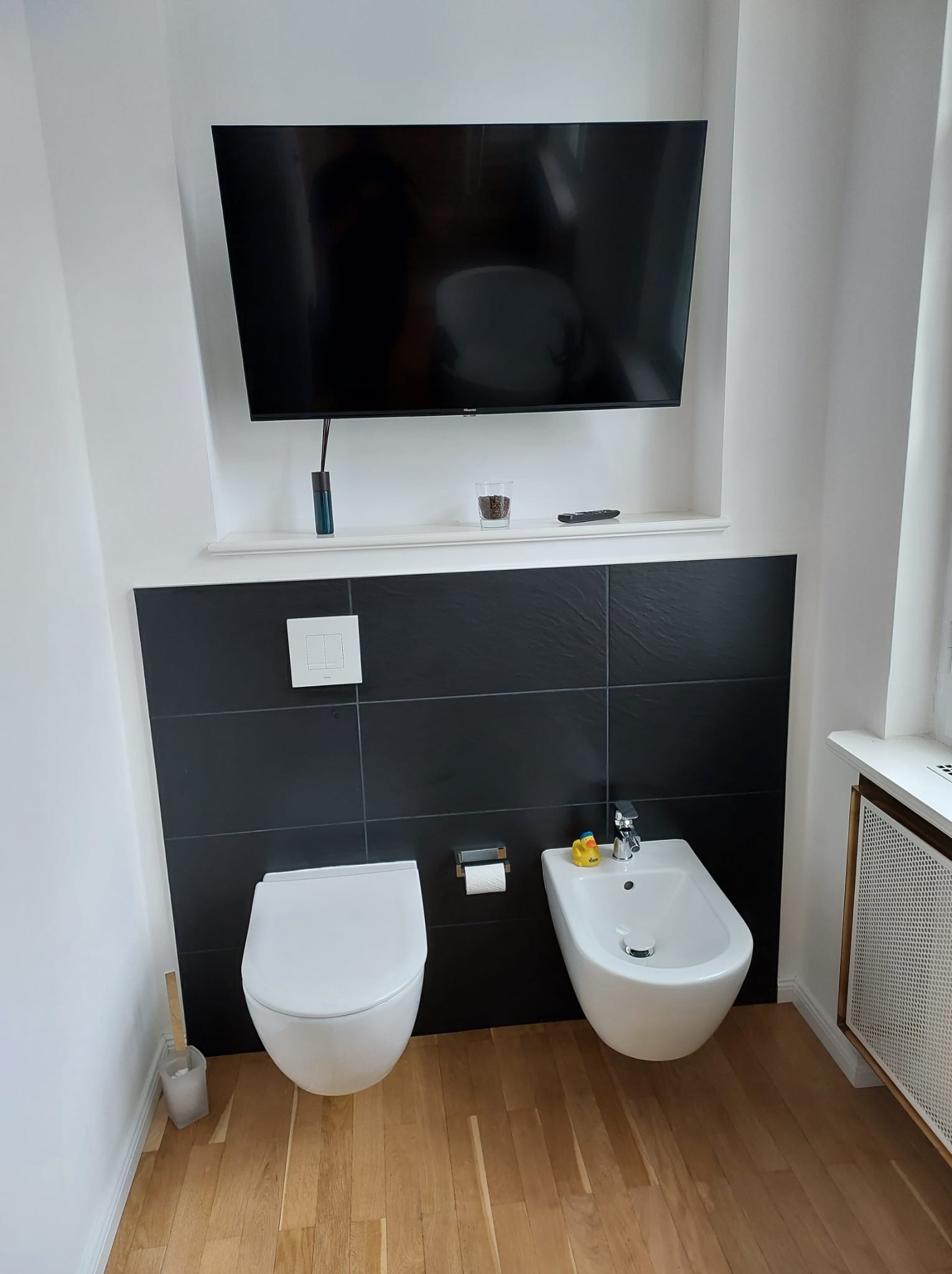Nowoczesna łazienka z podwieszaną toaletą i bidetem na tle czarnych płytek. Nad nimi półka z telewizorem. Na bidecie gumowa kaczuszka. Drewniana podłoga.