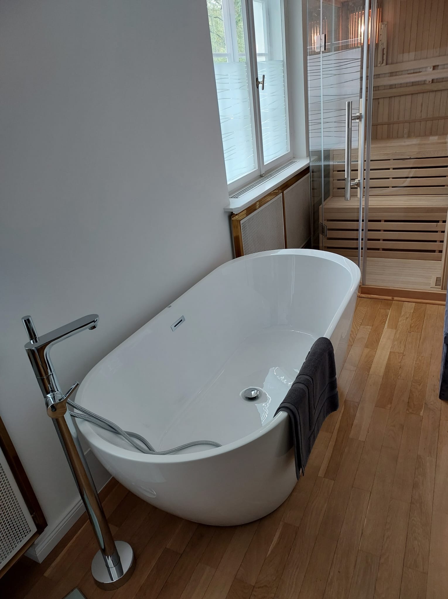 Nowoczesna łazienka z wolnostojącą wanną, chromowaną armaturą i drewnianą podłogą. W tle widoczna przeszklona sauna i okno z żaluzjami.