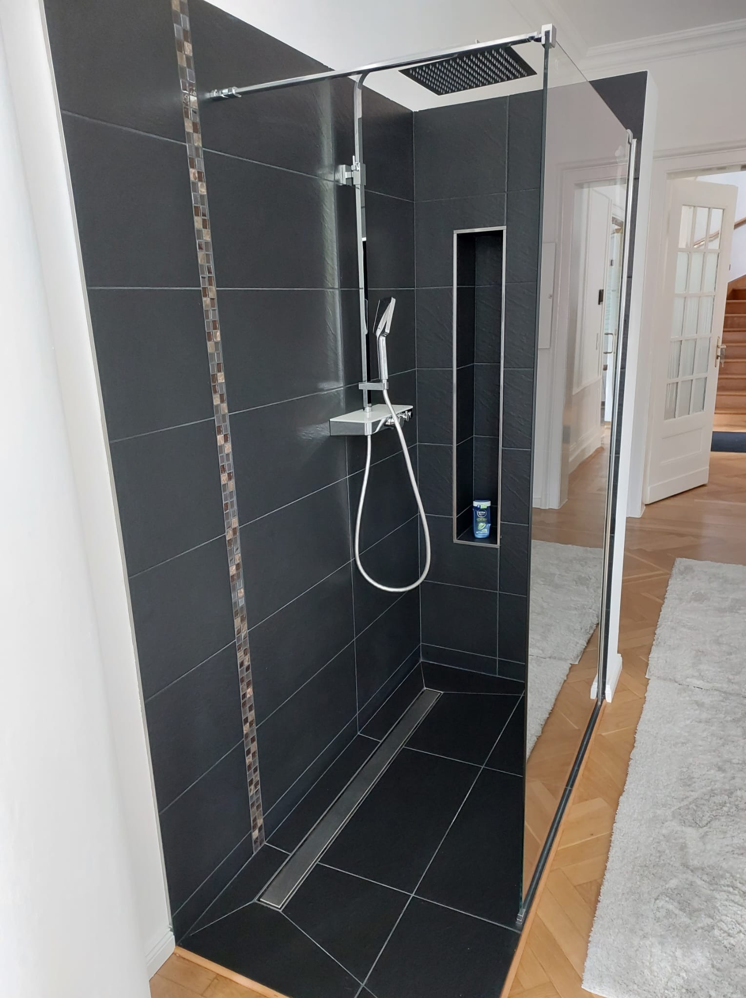 Nowoczesna kabina prysznicowa z czarnymi płytkami, mozaiką i szklaną ścianką. Widoczny odpływ liniowy i wnęka na kosmetyki. W tle parkiet i białe drzwi.