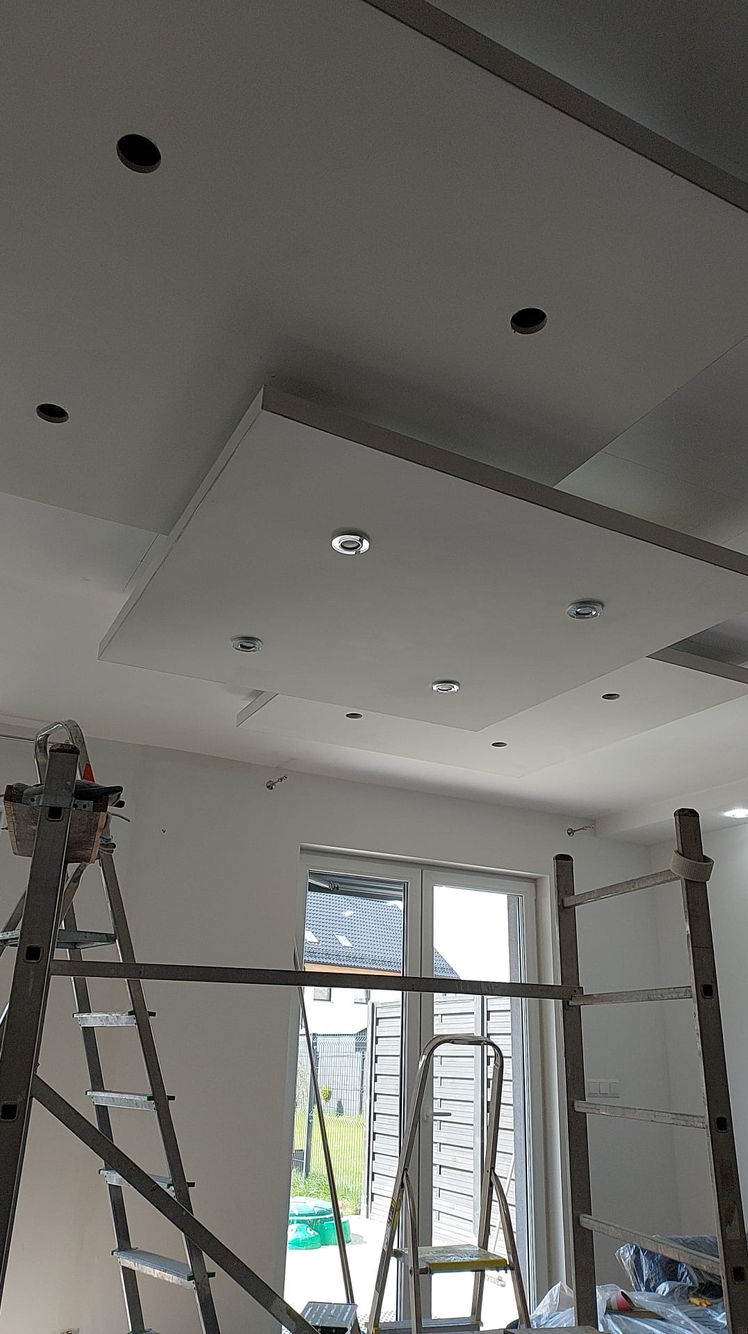 Biały sufit podwieszany z zamontowanymi punktowymi lampami LED, widok z dołu, drabiny malarskie w pomieszczeniu, okno w tle.