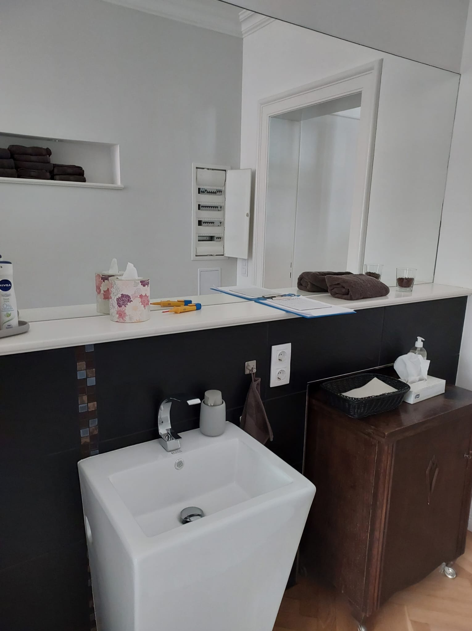 Nowoczesna łazienka po remoncie: biała, prostokątna umywalka, czarny pas z mozaiką, duże lustro, brązowe ręczniki i szafka w stylu retro. Widoczna otwarta szafka z bezpiecznikami.