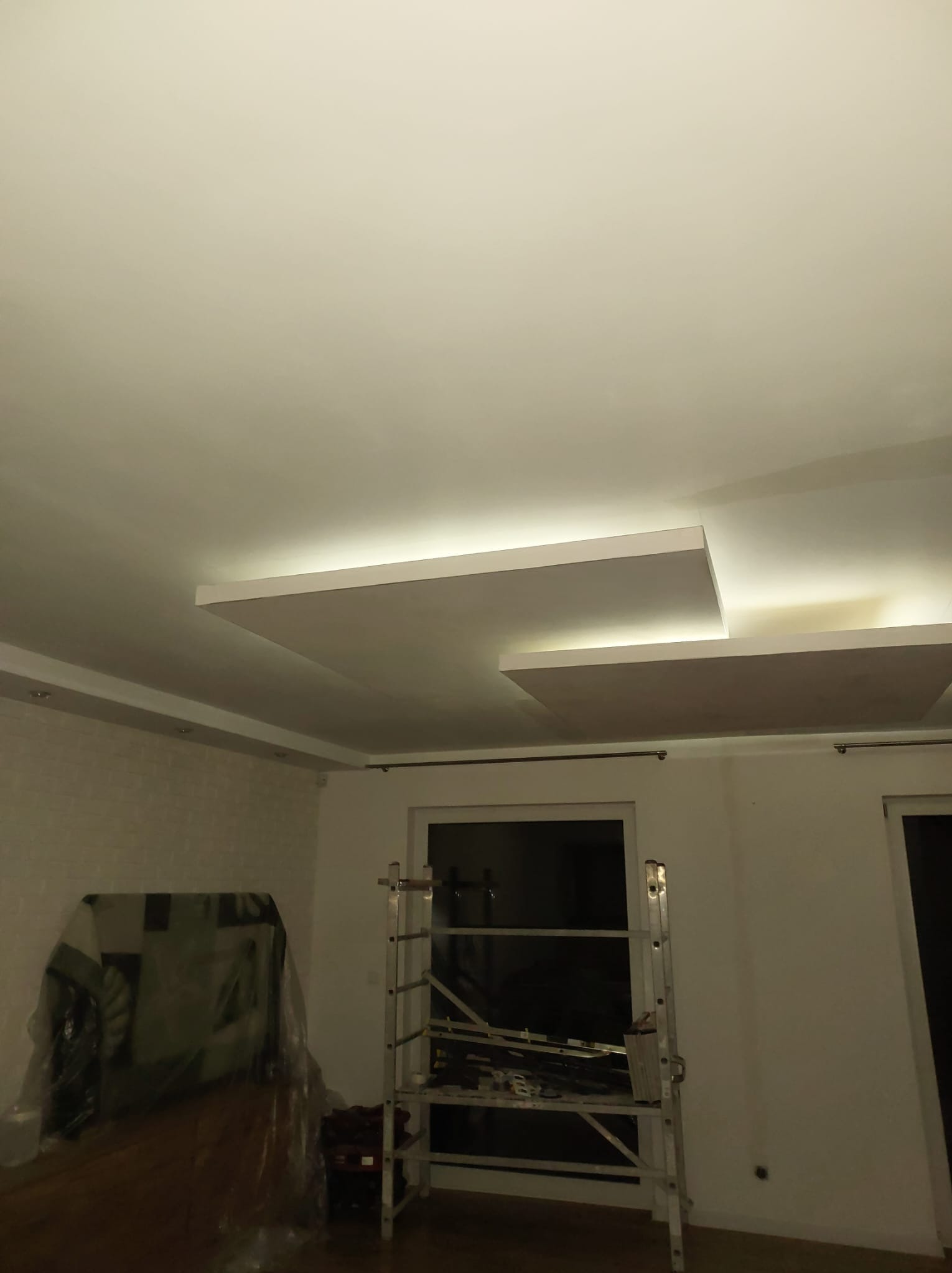 Nowoczesny sufit podwieszany z oświetleniem LED w trakcie montażu, widoczna drabina i zabezpieczone meble folią malarską. Neutralna kolorystyka wnętrza.