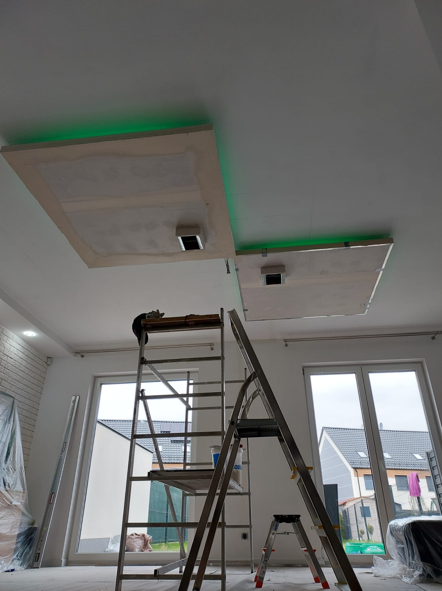 Sufit podwieszany z oświetleniem LED w trakcie montażu, widoczna drabina i narzędzia, surowy stan wykończenia, dwa okna w tle.