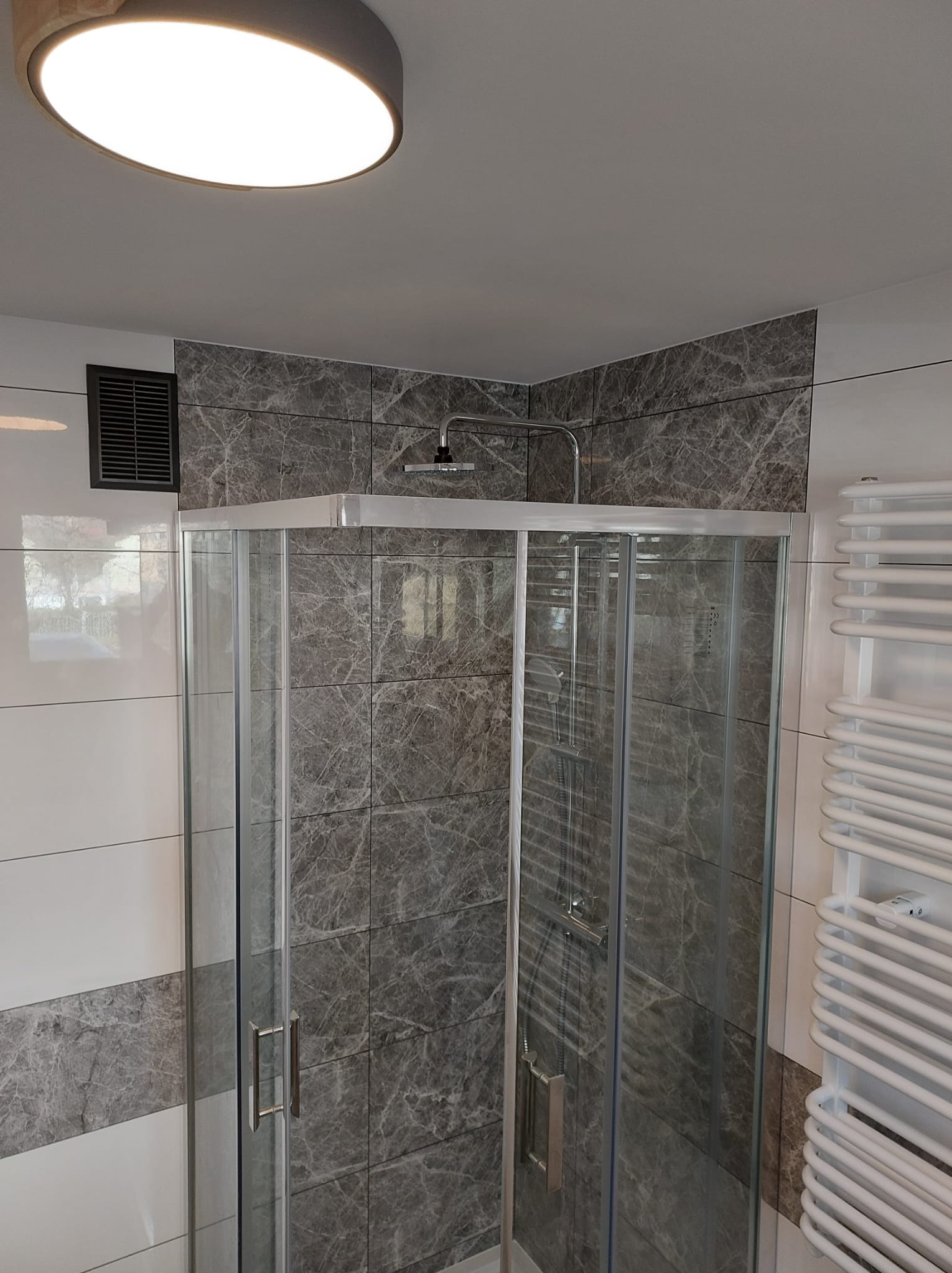 Nowoczesna kabina prysznicowa z szarymi płytkami imitującymi marmur, biały grzejnik drabinkowy i okrągła lampa sufitowa w wykończonej łazience.