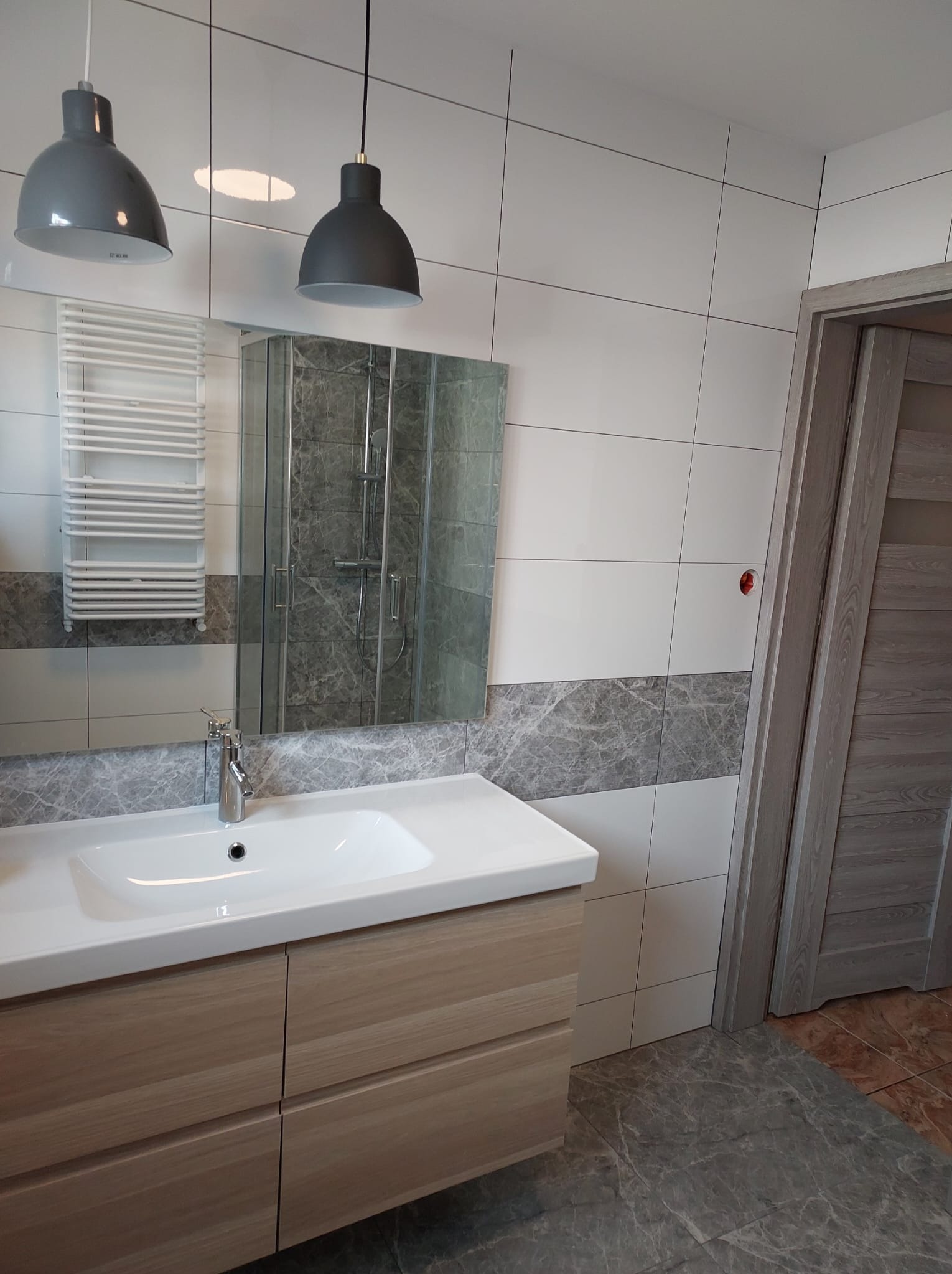 Nowoczesna łazienka z białymi i szarymi płytkami, drewnianą szafką pod umywalką i szklaną kabiną prysznicową. Dwie szare lampy wiszące nad umywalką.