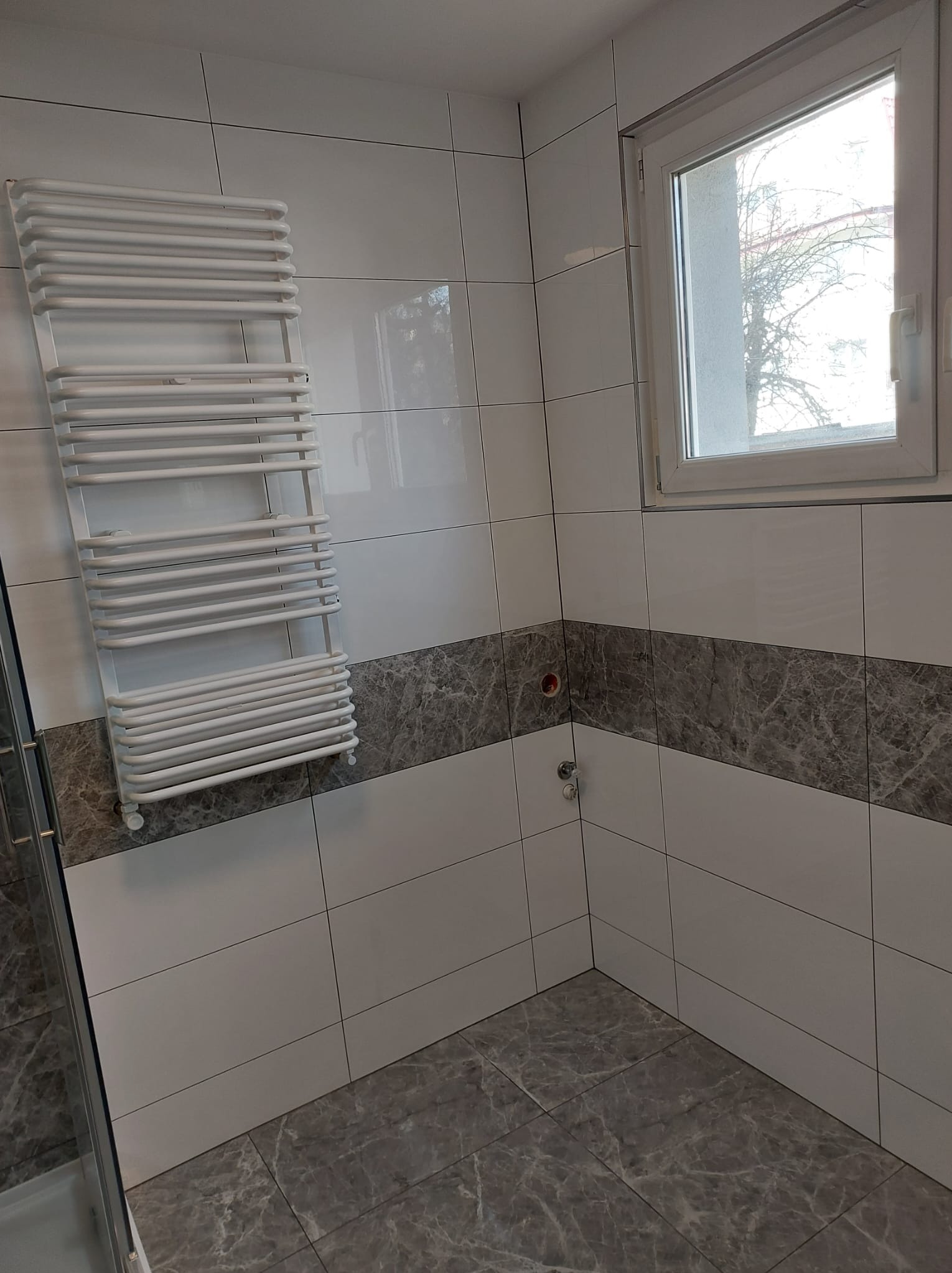 Wykończona łazienka z białymi i szarymi płytkami imitującymi marmur, grzejnikiem drabinkowym i oknem z widokiem na drzewo.