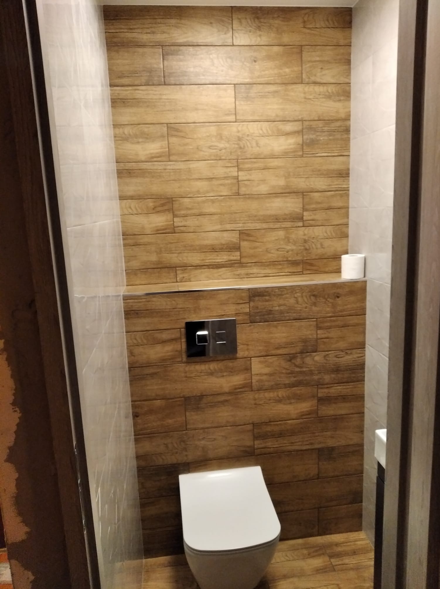 Nowoczesna toaleta z drewnopodobnymi płytkami na ścianie i podłodze, biała ceramika sanitarna, minimalistyczny design, widok z perspektywy drzwi.