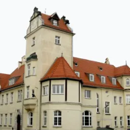 Odrestaurowany budynek z wieżą i nową, czerwoną dachówką, widoczny z perspektywy ulicy w pochmurny dzień. W prawym górnym rogu logo firmy dekarskiej.