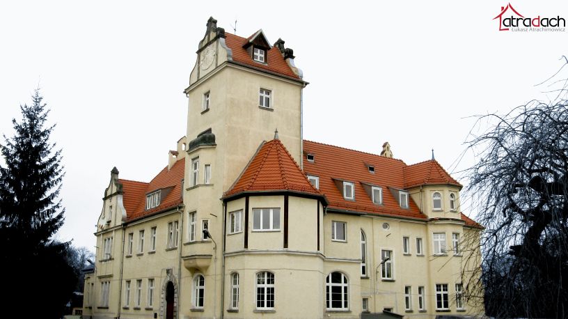 Odrestaurowany budynek z wieżą i nową, czerwoną dachówką, widoczny z perspektywy ulicy w pochmurny dzień. W prawym górnym rogu logo firmy dekarskiej.