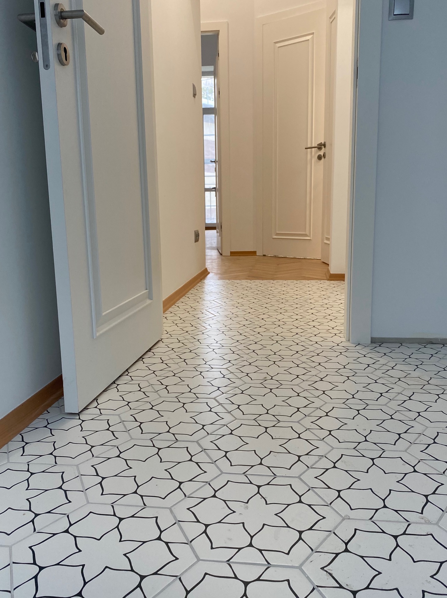 Biała podłoga z geometrycznym wzorem w korytarzu, widok na otwarte drzwi i fragment parkietu w tle. Listwy przypodłogowe w kolorze drewna.