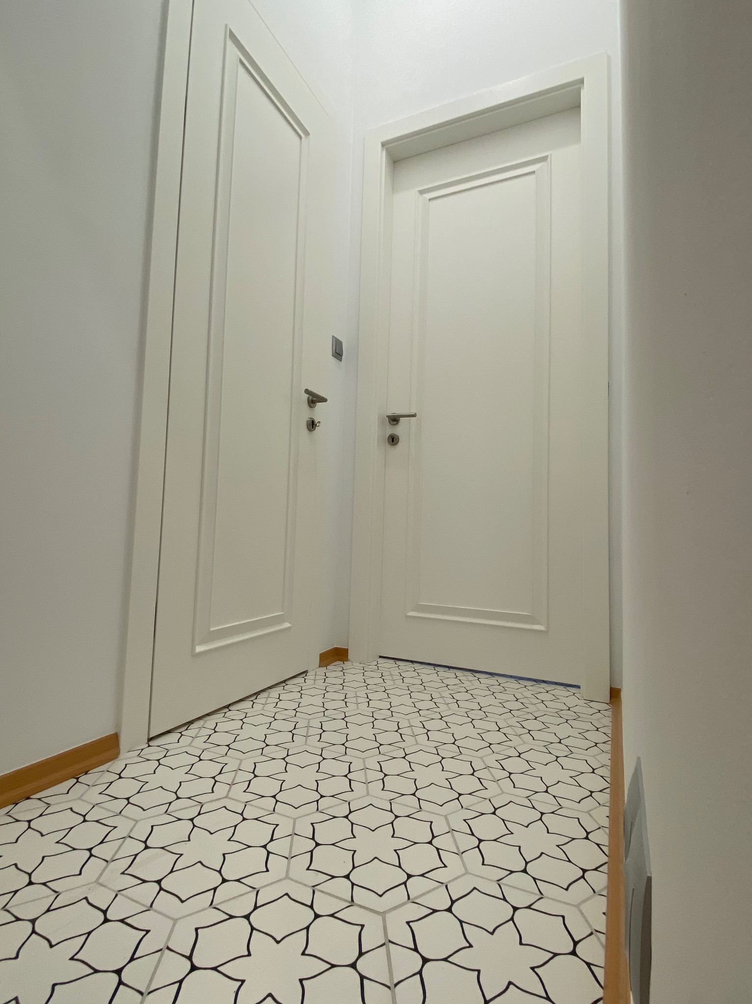 Białe drzwi wewnętrzne z ozdobnymi panelami i wzorzysta podłoga w odcieniach szarości tworzą nowoczesne wykończenie wnętrza. Widok z dołu podkreśla geometrię wzoru.