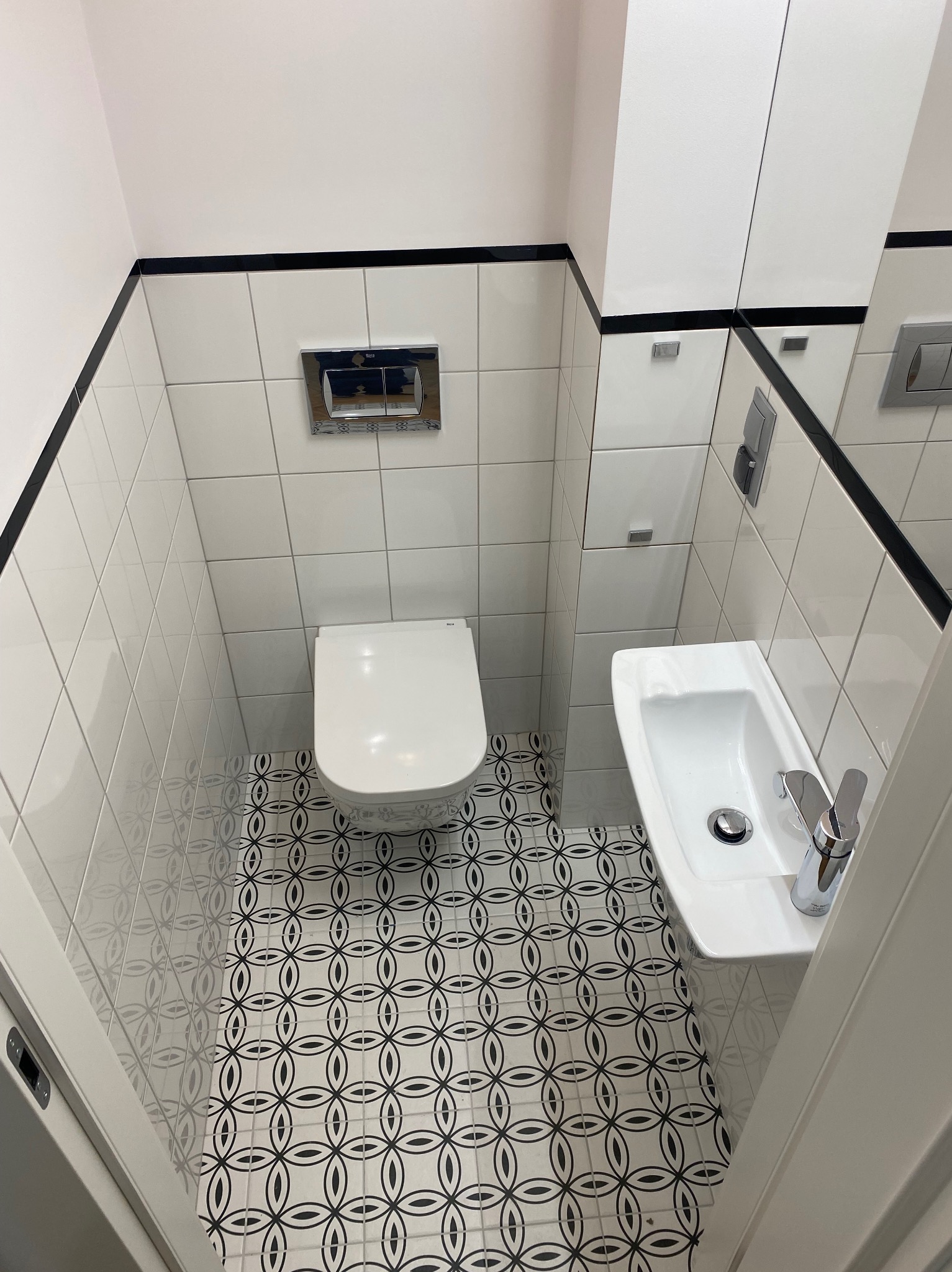 Kompaktowa łazienka z białą ceramiką, czarnymi akcentami i wzorzystą podłogą. Umywalka narożna, toaleta podwieszana, lustro z szafkami. Nowoczesny minimalizm.