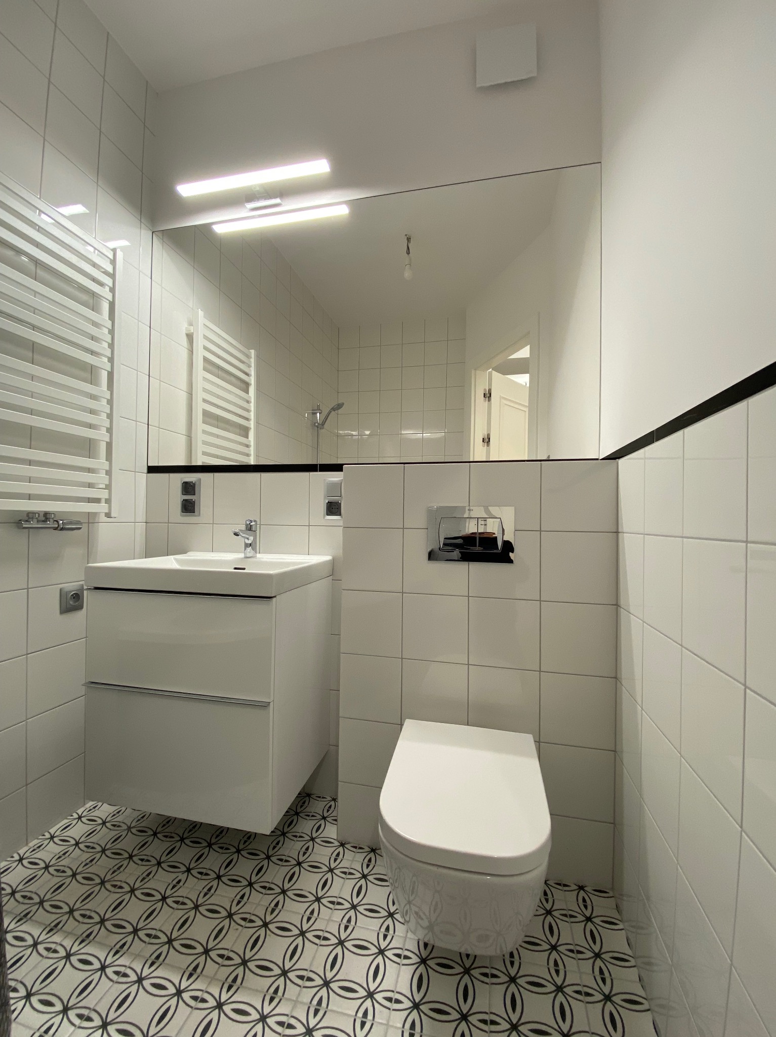 Nowoczesna łazienka z białymi płytkami, wzorzystą podłogą, wiszącą toaletą i umywalką z szafką. W lustrze odbija się fragment kabiny prysznicowej.
