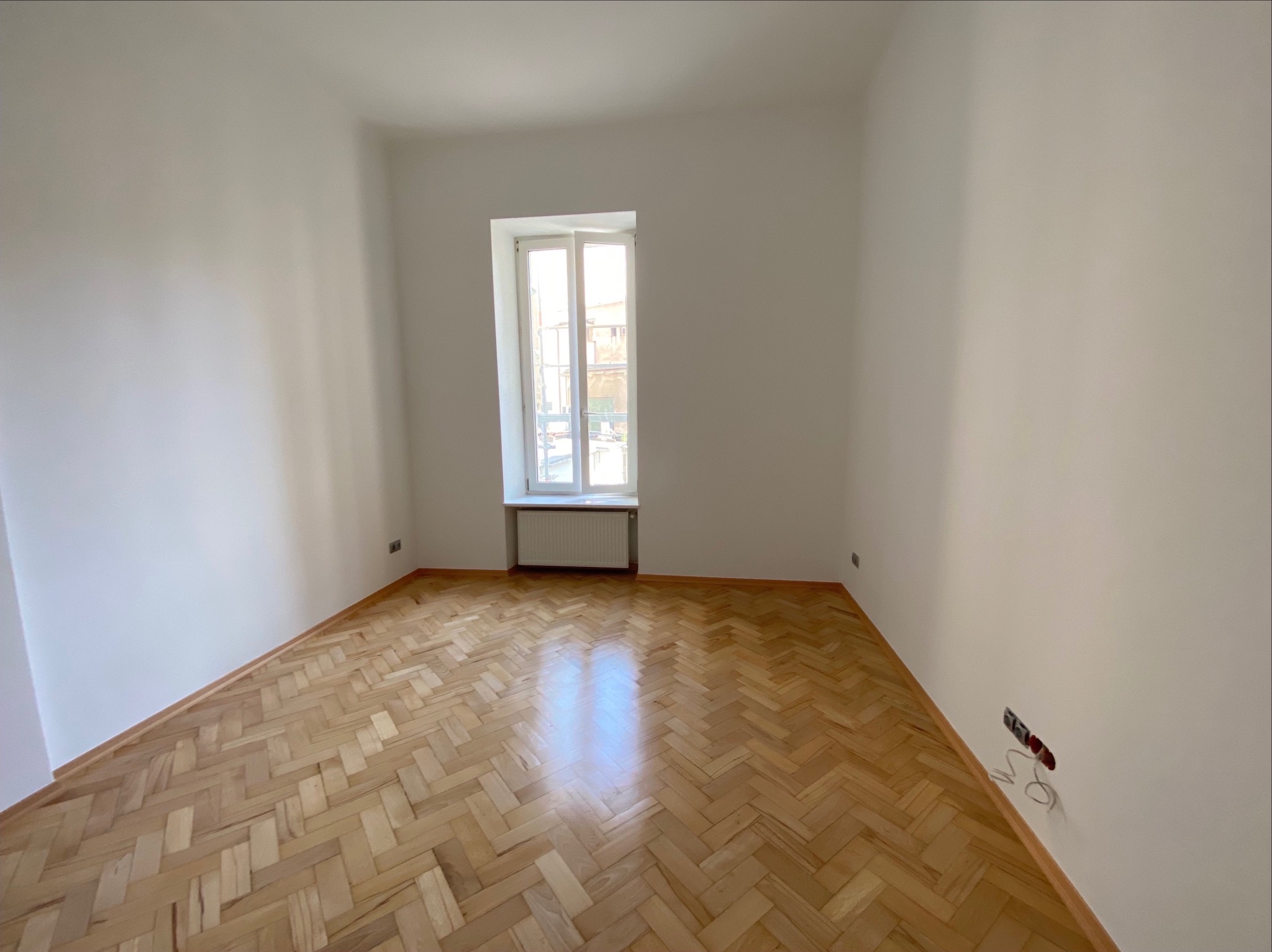Pusty pokój po remoncie z parkietem w jodełkę, świeżo pomalowanymi ścianami i oknem z grzejnikiem. Widoczne gniazdka elektryczne bez osprzętu.