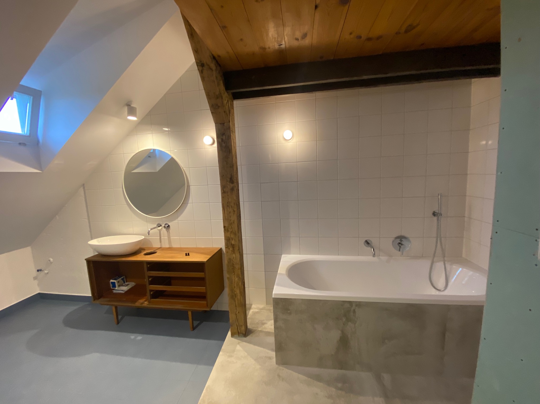 Jasna łazienka na poddaszu z drewnianą belką, białą wanną obudowaną betonem, umywalką na drewnianej szafce i niebieską podłogą. Ściany wyłożone białymi płytkami.
