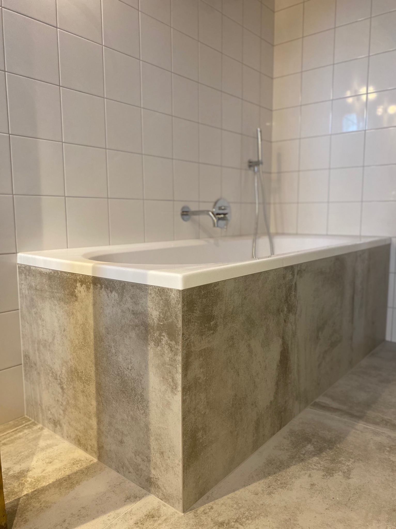 Nowoczesna wanna obudowana płytami imitującymi beton, białe płytki na ścianie, minimalistyczna armatura. Wykończona łazienka w neutralnej kolorystyce.