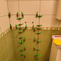 Marek Jan Śmietański - Ściana w łazience z zielonymi i beżowymi płytkami, na których zamontowano system poziomowania płytek z zielonymi klipsami i czerwonymi klinami. Widoczny fragment toalety i narzędzia.