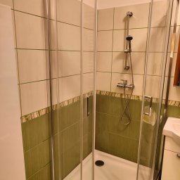 Marek Jan Śmietański - Kabina prysznicowa narożna z chromowanymi profilami i transparentnym szkłem, widoczna armatura i zielono-beżowe płytki w łazience. Umywalka z szafką po prawej.