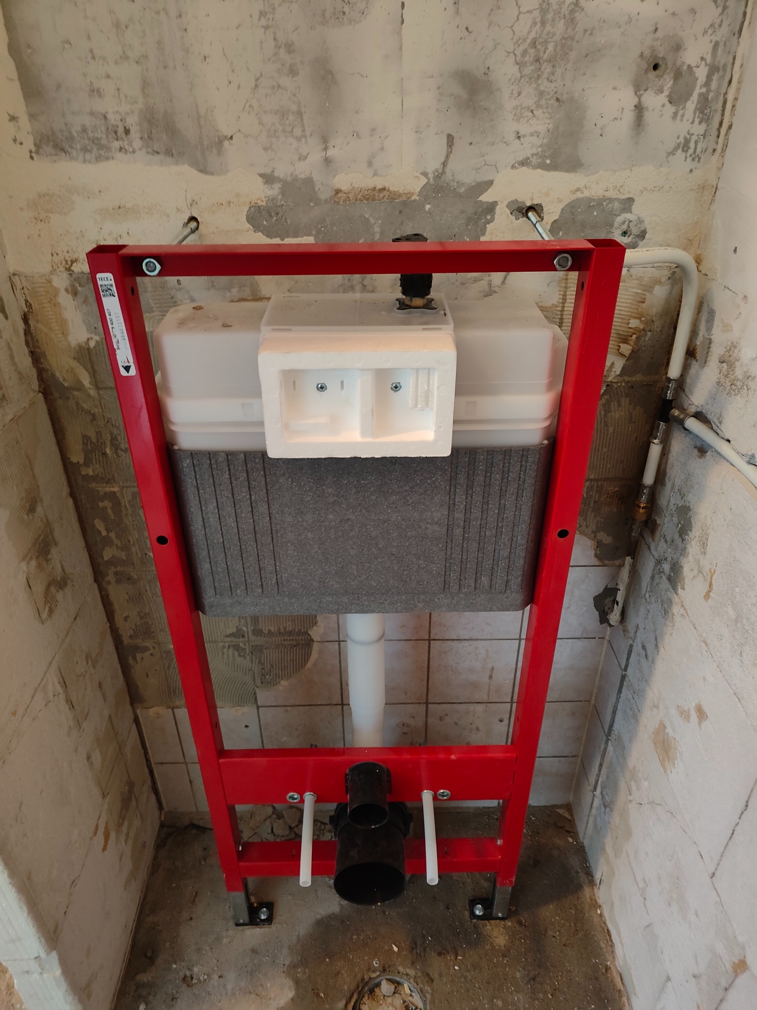 Czerwony stelaż podtynkowy WC zamontowany w surowej łazience, widoczne rury instalacyjne i fragmenty starej glazury na ścianach. Ujęcie z dołu.